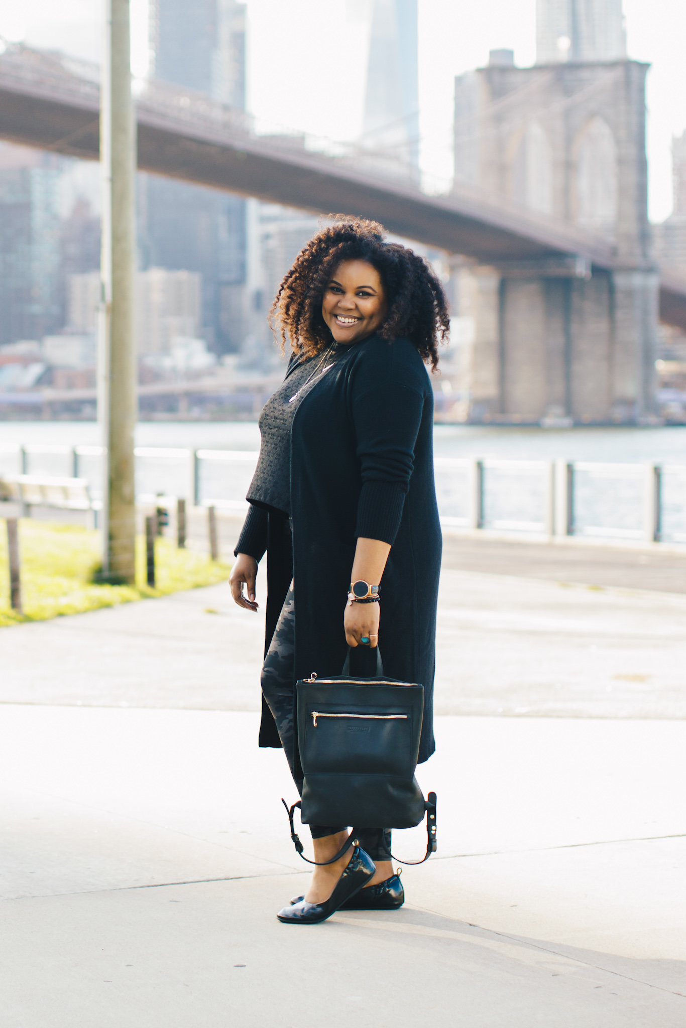 black woman wears long black cardigan holding a black leather backpack