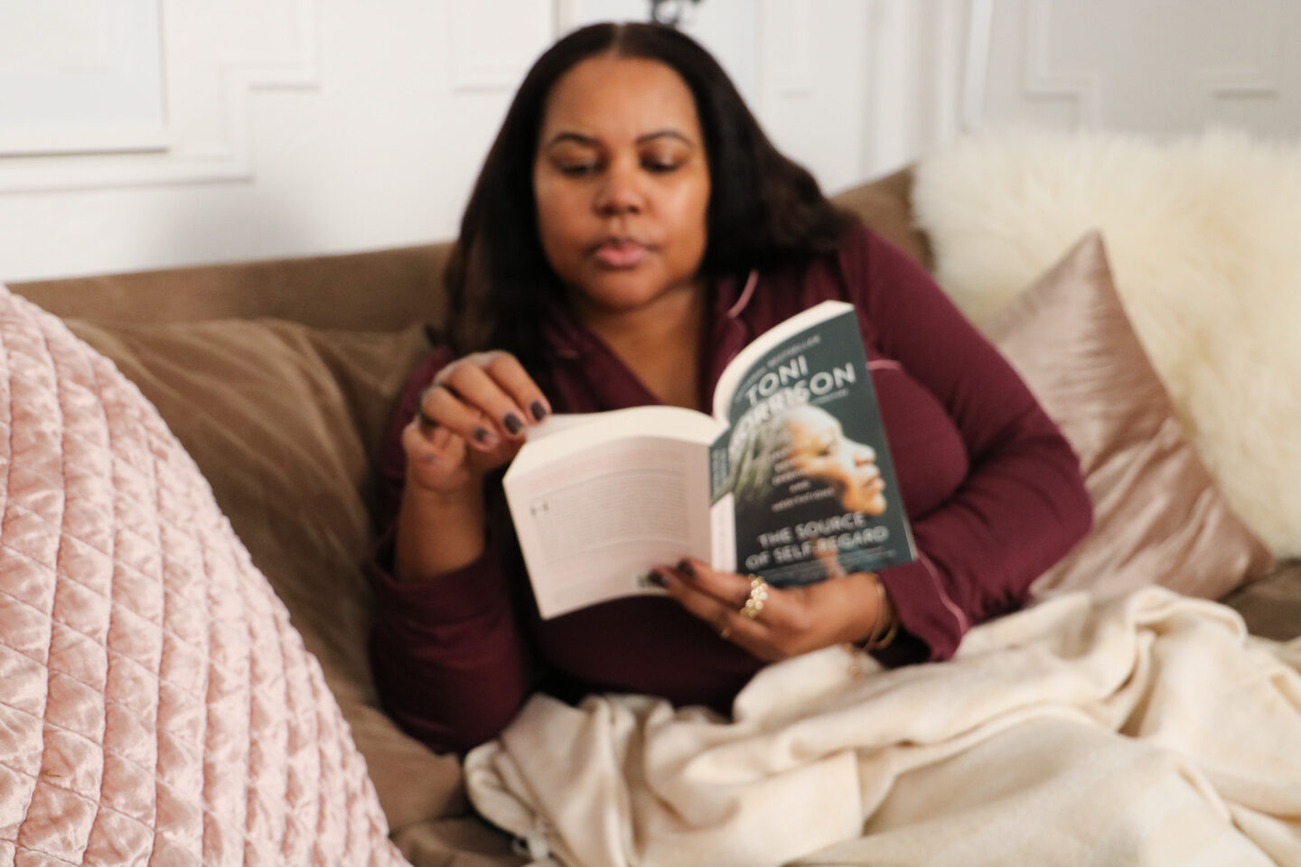 Patranila sits on a sofa reading The Source of Self Regard by Toni Morrison